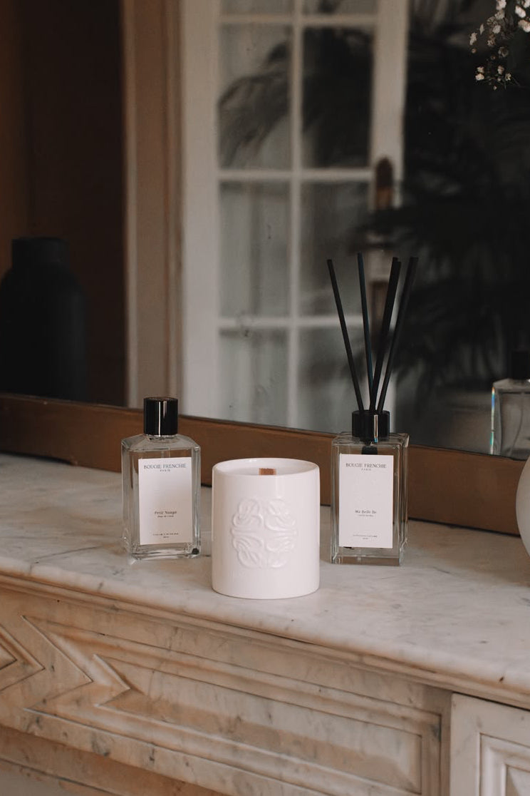 Une main allume une bougie parfumée Bougie Frenchie, à côté du parfum d'intérieur Petit Nuage